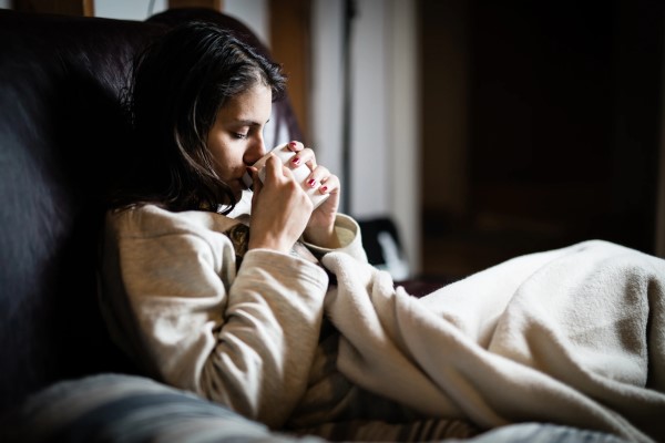 Woman drinking mug of hot tea on a sofa under a fleece blanket.