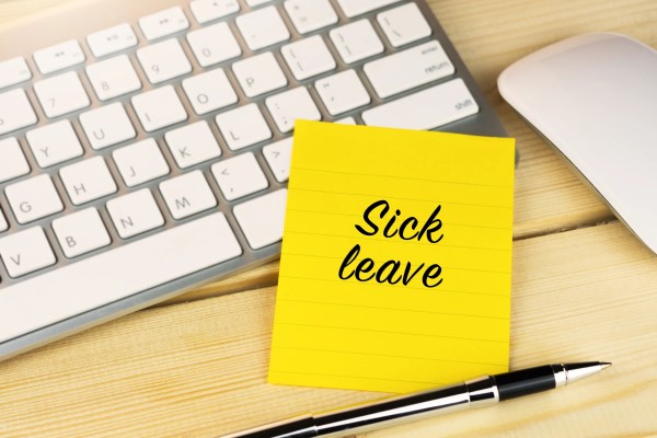 Yellow post-it note with the words "Sick leave" attached to an office desk