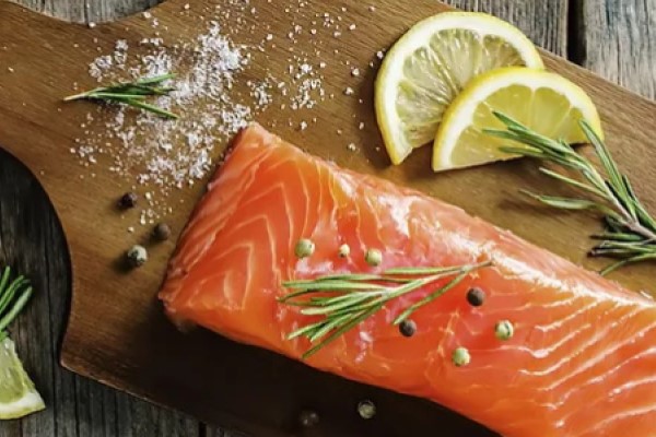 Piece of raw, fresh salmon on a cutting board with rosemary, peppercorns, salt, and lemon wedges