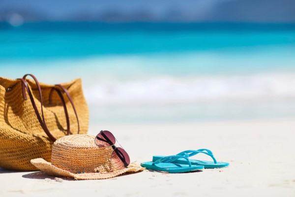 Woven bag, woven hat, sunglasses, and flip flops lying on the beach near pristine blue water