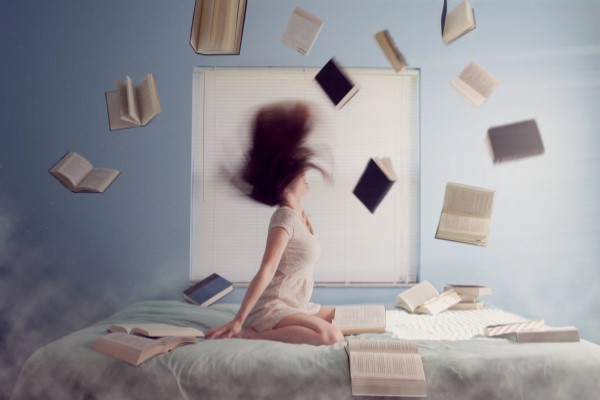 Woman on bed flipping her hair back, some books flying in the air around her and some books laying on the bed around her
