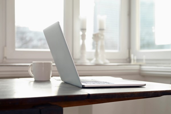 Laptop and white mug sitting on a wooden table surrounded by large windows