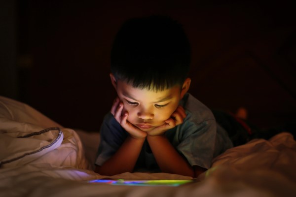Young boy laying on a bed in the dark staring at a digital tablet screen