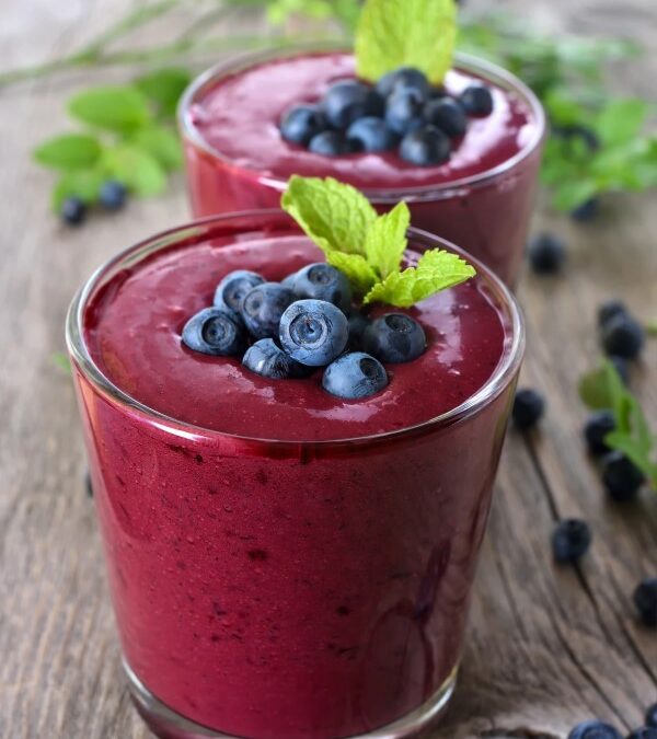Two glasses filled with dark purple smoothies and topped with fresh blueberries and mint leaves