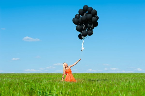 Woman standing in tall, green grass letting a bunch of black balloons float off into a cloudless sky