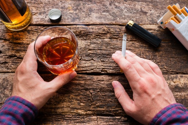 Hands holding a lit cigarette and a rocks glass of amber alcohol