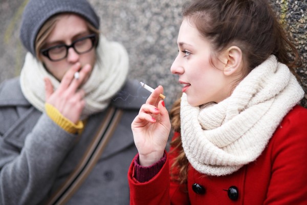 Two people standing outside, bundled up, smoking cigarettes